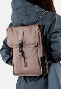 Zaino in nylon rosa con parte superiore a patta, tracolla regolabile, hardware nero e superficie testurizzata. Presenta un grande scomparto principale e una tasca laterale.