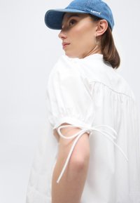 Γυναίκα που φοράει λευκό μπλουζάκι με φουσκωτά μανίκια και δεμένες μανσέτες, ασημένια σκουλαρίκια κρίκους και μπλε καπέλο μπέιζμπολ της Levi's, κοιτάζοντας πάνω από τον ώμο της.