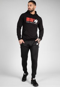Hoodie noir avec "GORILLA WEAR" et "ÉTATS-UNIS D'AMÉRIQUE" en texte rouge et blanc. Assorti à un jogger noir avec des fermetures éclair et un logo.
