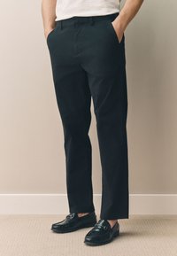 Chinos noirs à coupe slim, dotés de poches latérales et d'une braguette zippée, associés à des mocassins noirs brillants sur un fond neutre.