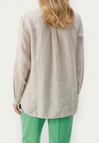 Lysebeige langærmet skjorte med løs pasform, med rygplissé og knapmanchetter, kombineret med grønne bukser. Glat, tekstureret stof.