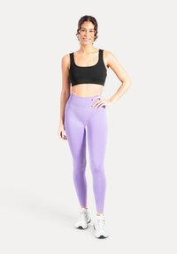 Schwarzer Sport-BH mit breiten Trägern und kurzem Schnitt, kombiniert mit hoch taillierten, enganliegenden lilafarbenen Leggings. Das Outfit wird mit Turnschuhen komplettiert.