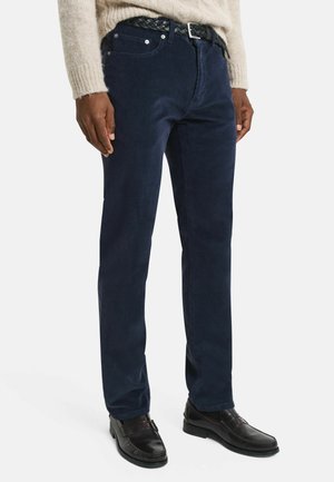 Man wearing dark blue corduroy pants with black braided belt, beige sweater, and black leather loafers on white background.