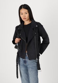 Black faux suede motorcycle jacket with a notched collar, silver zippers, side pockets, and adjustable belt detailing. Paired with light jeans.