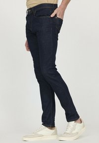 Jeans skinny en denim bleu foncé avec des coutures orange. Comprend une poche avant et une fermeture éclair, associés à des baskets crème.