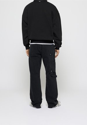 Person trägt schwarze Cargohose und schwarzen Pullover, steht mit dem Rücken zur Kamera auf einfarbigem Hintergrund.