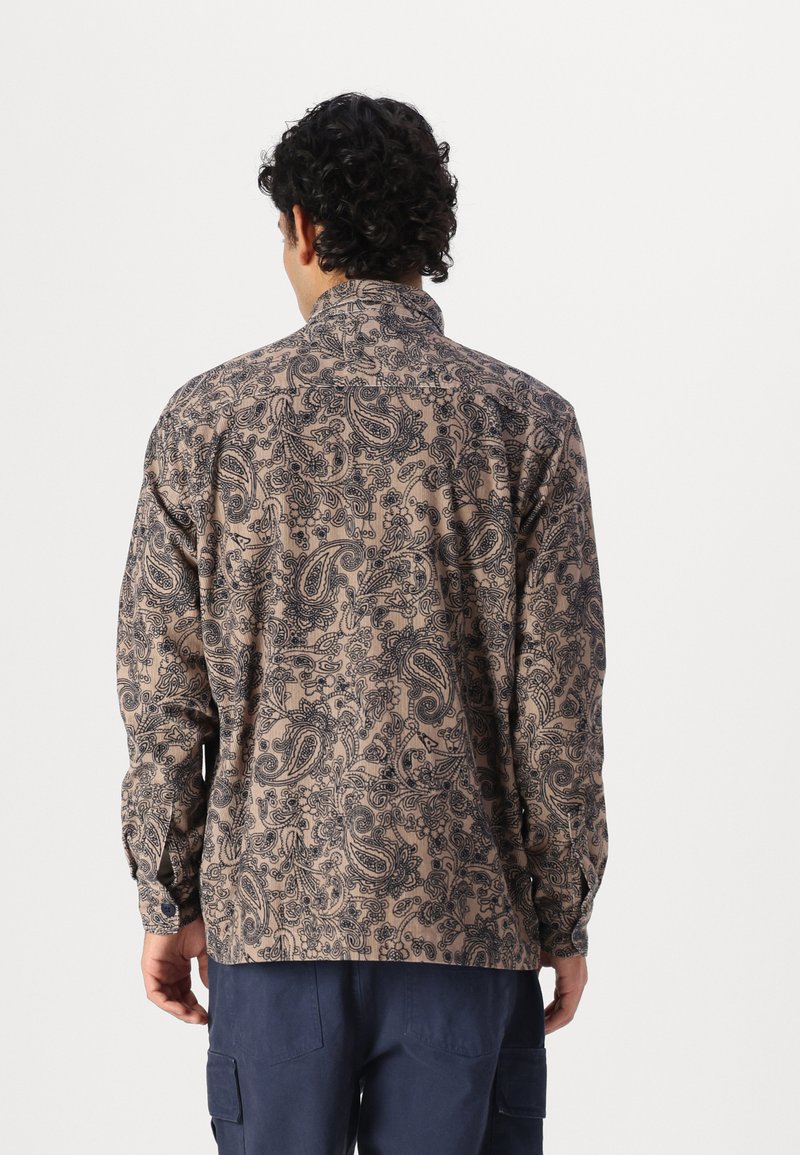 Chemise beige à boutons, avec un motif paisley noir, manches longues et col, portée à l'envers, mettant en valeur le design et la texture délicats.