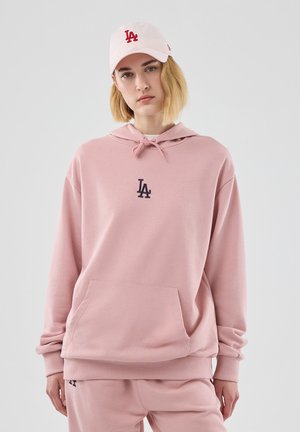 Mujer que lleva una sudadera con capucha rosa pálido y pantalones a juego con el logo "LA", y una gorra rosa clara con un logo rojo "LA", de pie frente a un fondo neutro.
