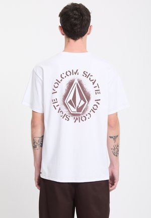 Jeune homme aux cheveux courts et bruns portant un T-shirt blanc avec un logo circulaire Volcom Skate marron dans le dos et un pantalon foncé.