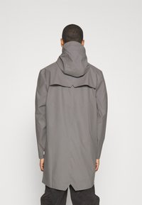 Grå vattentålig jacka med hög krage, långa ärmar och en detaljerad vent bak. Har en huva och en smal, rak design.