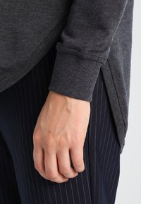 Maglia grigia a maniche lunghe con texture a coste e spacchi laterali, abbinata a pantaloni navali a righe. Primo piano di una mano appoggiata sul tessuto.