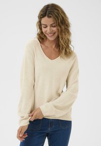Lächelnde Frau mit welligem Haar, die einen cremigen Strickpullover und eine blaue Jeans trägt und den Ärmel anpasst, mit einem schlichten weißen Hintergrund.