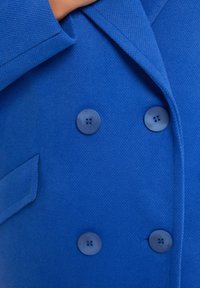 Dwurzędowa niebieska marynarka wykonana z teksturowanej tkaniny. Posiada płaskie klapy oraz okrągłe, ciemnoniebieskie guziki z czterema otworami.