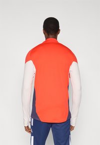 Rød og hvit langermet treningsskjorte med marineblå detaljer på sidene, med tettsittende design og glatt tekstur.