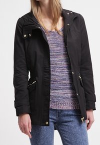 Black rain jacket with a removable hood, zippered front, two side pockets, and gold-tone hardware, layered over a purple knit sweater.