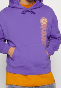 Violetti huppari, jossa on etutasku ja oranssi graafinen suunnittelu vasemmalla puolella. Vaatteen päällä on oranssi paita.