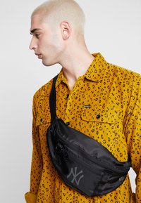 Sacoche en nylon noir au design épuré avec un logo de New York. L'utilisateur porte une chemise jaune à motif léopard.
