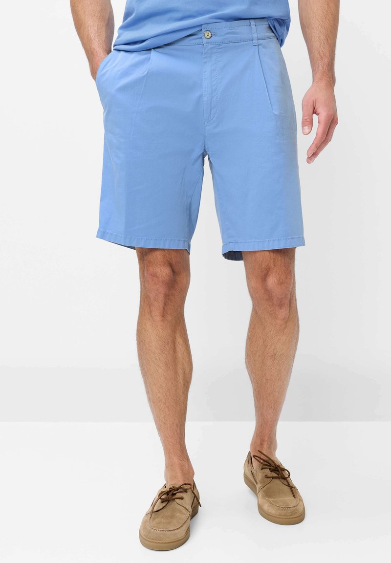Mann trägt hellblaue Shorts mit Seitentaschen und hellbraune Schnür-Bootsschuhe, steht vor einem schlichten weißen Hintergrund.