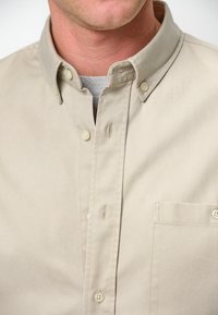 Camicia beige con bottoni, dotata di colletto classico, con taschino sul petto e piccoli bottoni color crema. Realizzata in tessuto morbido e texture.