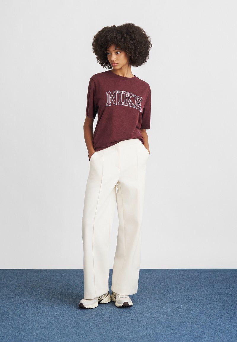 Maronenfarbenes Nike T-Shirt mit kurzen Ärmeln und großem Logo, kombiniert mit hochgeschnittenen weißen Hosen. Das Modell trägt hellfarbene Sneakers.