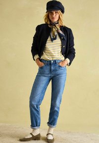 Schwarze strukturierte Jacke, gestreiftes gelb-weißes Hemd, blaue hochtaillierte Jeans, gepunktete Socken, dunkle Schuhe und eine dunkle Mütze.