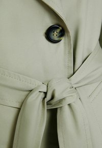 Světlý zelený trenčkot z látky s velkým černým knoflíkem, stahovacími pásky v pase a viditelnými detailem šití.