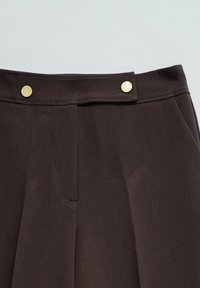 Pantalones marrón oscuro en la zona de la cintura con dos cierres de botón en tono dorado en una solapa de cintura extendida.