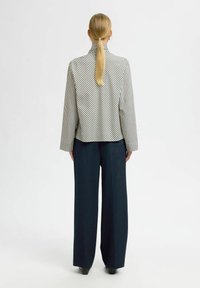 Kvinde med lang blond hestehale iført en hvid og sort stribet blazer og brede navy bukser, stående foran en ensfarvet hvid baggrund.