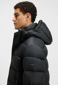 Chaqueta puffer negra con capucha, que presenta secciones acolchadas, un collar alto y un parche de marca en la mangas izquierda. Textura suave.