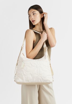 Giovane donna con top verde oliva senza maniche e pantaloni beige che tiene una grande borsa a tracolla bianca trapuntata, guarda di lato su sfondo uniforme.