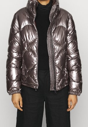 Persoon die een glanzend metallic paarse pufferjas met een hoge kraag draagt over een zwarte gebreide trui en zwarte broek.