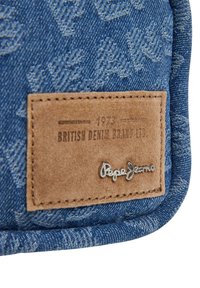 Mėlyna džinsų audinys su tekstūriniu raštu, kuriame yra šviesiai rudos odos etiketė, išlakuota užrašu "British Denim Brand Ltd" ir sidabro "Pepe Jeans" logotipas.