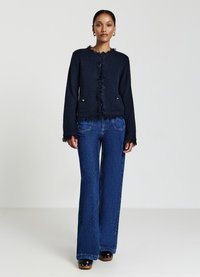 Marineblauer strukturiert Cardigan mit Fransen, mit goldfarbenen Akzenten und Taschen. Kombiniert mit weit geschnittenen blauen Jeans.