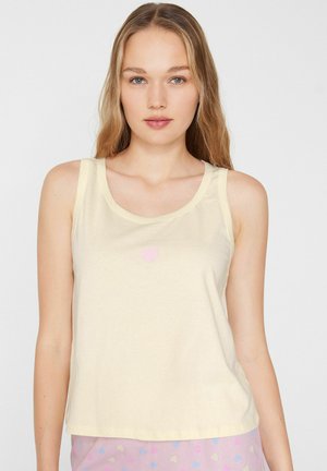 Jeune femme aux longs cheveux blonds portant un top sans manches jaune clair avec un petit cœur rose et un pantalon à motifs cœurs pastel.
