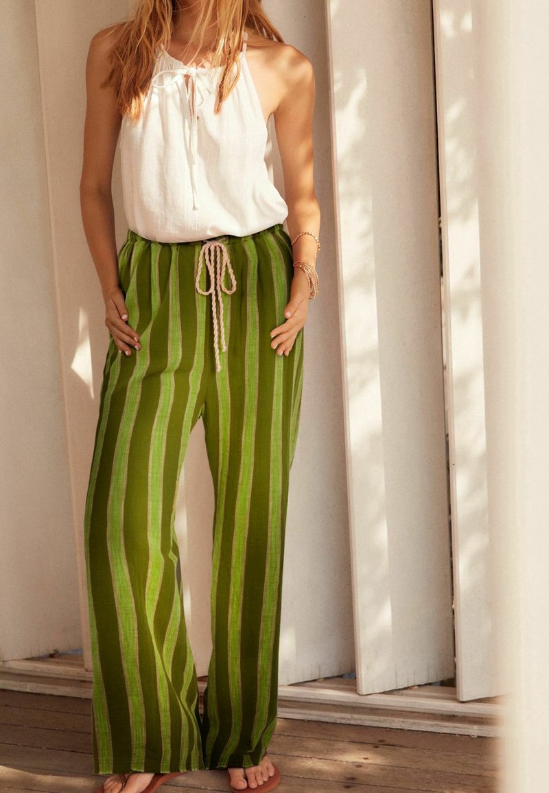 Femme portant un haut blanc sans manches et un pantalon ample vert rayé, debout pieds nus sur un sol en bois contre un mur de couleur claire.