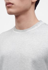 Gros plan sur le cou et la partie basse du visage d’un homme portant un t-shirt gris clair à col rond avec le texte "MICHAEL KORS" sur le col.