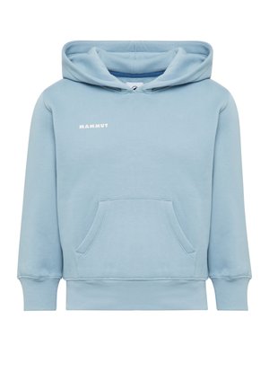 Un ragazzo giovane è in piedi con una felpa con cappuccio azzurro chiaro, pantaloni giallo brillante, scarpe da ginnastica bianche e porta uno zaino blu navy su una spalla davanti a uno sfondo semplice.