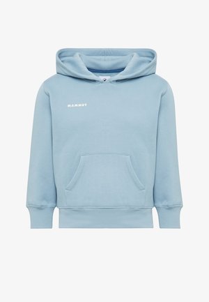 Un jeune garçon se tient debout portant un sweat à capuche bleu clair, un pantalon jaune vif, des baskets blanches et un sac à dos bleu marine sur une épaule devant un fond uni.