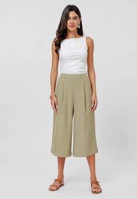 Bílý upnutý top bez rukávů v kombinaci s kalhotami s vysokým pasem, širokými nohavicemi a světlou khaki barvou. Model má na sobě hnědé ploché sandály a kruhové náušnice. Hladké textury.