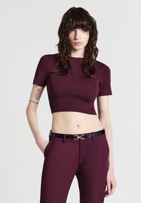 Una giovane donna con lunghi capelli scuri indossa un top corto attillato bordeaux e pantaloni abbinati con una cintura nera, in piedi contro uno sfondo semplice.