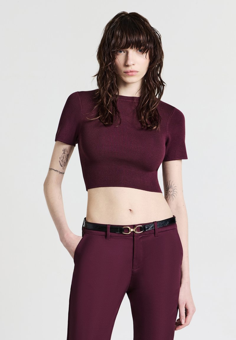 Una giovane donna con lunghi capelli scuri indossa un top corto attillato bordeaux e pantaloni abbinati con una cintura nera, in piedi contro uno sfondo semplice.