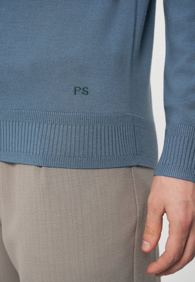 Jasnoniebieski sweter z prążkowanymi mankietami i dolnym obszyciem, z zielonym haftem "PS"; zestawiony z teksturowanymi beżowymi spodniami.