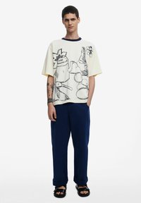 Light-colored t-shirt with large black line drawings and a ribbed navy collar, paired with navy loose-fitting trousers and black sandals.