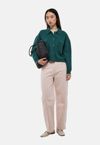 Cardigan en tricot vert avec deux poches poitrine, pantalon large rose pâle, et sac à main noir en osier. Des ballerines beiges complètent l'ensemble.