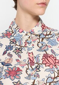 Bluse mit floralem Muster in Creme mit blauen und pinken Blumen. Verfügt über eine Knopfleiste und einen spitzen Kragen. Aus leichtem Stoff hergestellt.