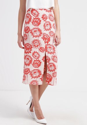 La mujer lleva una falda blanca hasta la rodilla con grandes estampados florales rojos y una apertura frontal, combinada con zapatos blancos de tacón alto.