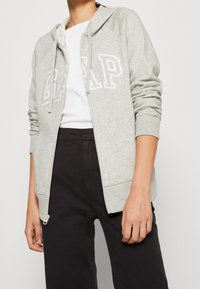 Sudadera gris con cremallera y logo blanco "GAP", con capucha con cordón, puños acanalados y bolsillos delanteros, combinada con pantalones negros.