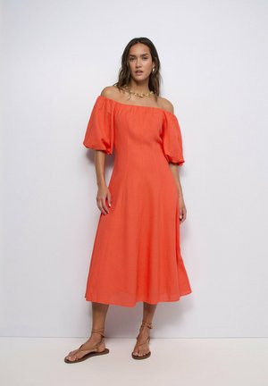 Mujer con un vestido midi naranja brillante con hombros descubiertos y mangas abullonadas, y sandalias planas con tiras, sobre un fondo blanco liso.