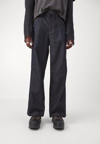 Dark blue denim pants with a high waist, wide leg design, and visible yellow stitching. Paired with black shoes and a charcoal shirt.