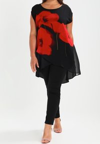 Černá volná tunika s velkým květinovým potiskem červených máků, spárovaná s černými přiléhavými kalhotami a nahými lodičkami. Hladká textura a zakřivený lem.
